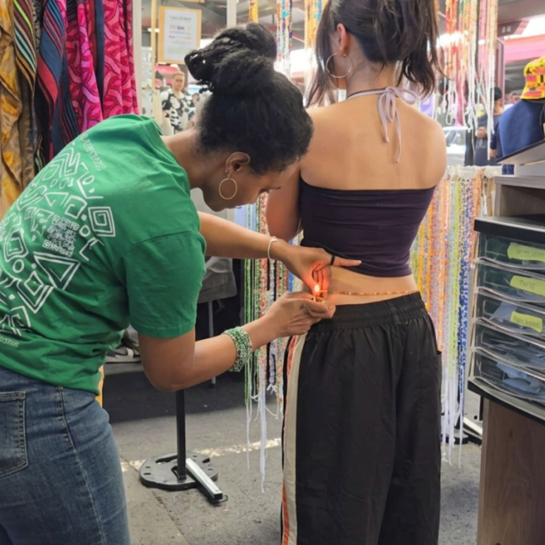Person getting waistbeads tied on their waist at at a market stall with colourful waistbeads in the background.